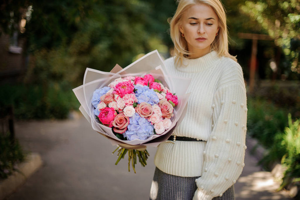 Girl in the white sweather holding in her hands a beautiful bouquet of tender pink and blue autumn flowers in white transparent paper - Фото, изображение