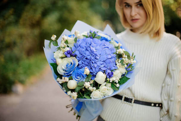Blonde girl in the white sweather holding in her hands bouquet of tender blue flowers in blue paper - Фото, зображення