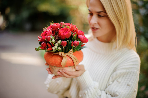 Blonde girl in the white sweather holding in her hands a pumpkin decorated with red flowers with red hot peppers and green leaves - Foto, afbeelding