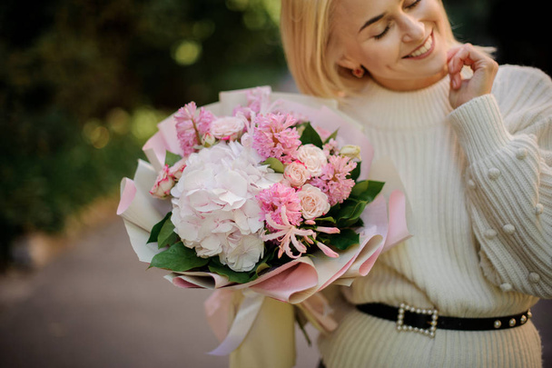 Smiling girl in the white sweather holding in her hands a huge bouquet of tender pink flowers decorated with green leaves - Fotografie, Obrázek