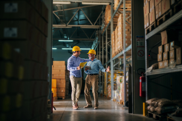 Picture of two business man standing in warehouse with helmets on their heads and celebrating great news about their business. Standing in big warehouse and looking happy and satisfied. - Photo, Image