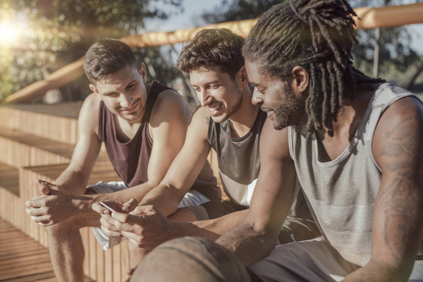 Grupo multiétnico de jugadores de baloncesto sentados junto a la cancha y utilizando el teléfono celular
. - Foto, imagen