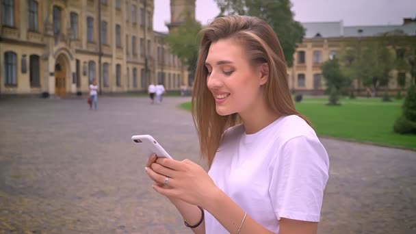 awesome caucasian girl is scrolling her phone while standing in profile on the street chill and focused with smirk, urban view - Кадри, відео