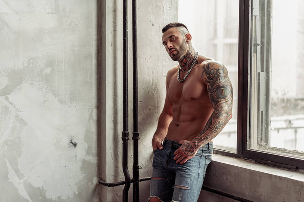 Fashion portrait of Sexy naked male model with tattoo and a black beard standing in hot pose on near the window. Loft room interior with grey concrete wall. Professional Studio image. - Zdjęcie, obraz