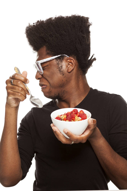 angeekelter junger gutaussehender afroamerikanischer Hipster, der Müsli mit frischen Früchten isst, isoliert auf weißem Hintergrund. gesundes Ernährungskonzept - Foto, Bild