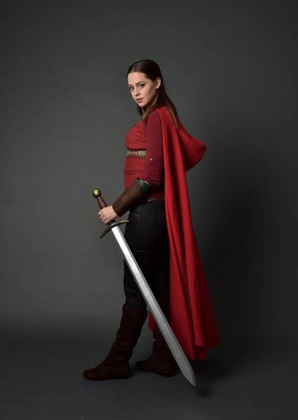 po celé délce portrét bruneta dívka, která nosí červený středověký kostým a plášť. stojící pozice drží meč na šedé studio pozadí. - Fotografie, Obrázek