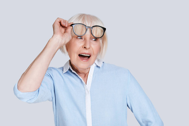 mulher caucasiana envelhecida chocada com boca aberta posando em fundo cinza no estúdio e ajustando óculos elegantes
  - Foto, Imagem