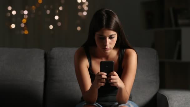 Front view of a sad girl reading cyber bullying phone message sitting on a couch in the night at home - Záběry, video