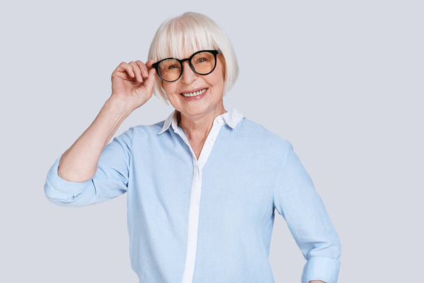 aged caucasian woman with short grey hair posing at grey background in studio and adjusting stylish glasses  - Фото, зображення