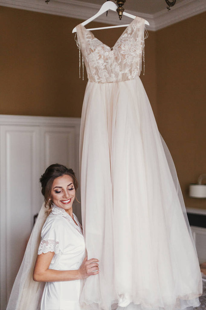 Gorgeous bride in silk robe looking at stylish wedding dress, hanging on chandelier in room in the morning.Happy Bride and her beautiful wedding gown. Woman getting ready - Zdjęcie, obraz