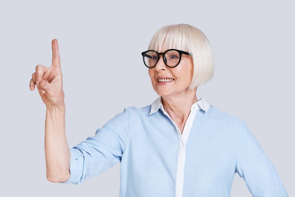 smiling stylish aged stylish woman with short grey hair posing at grey background in studio and pointing finger up - Foto, immagini