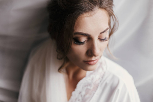 Gorgeous beautiful bride in silk robe posing near window in soft light in the morning. Happy stylish Bride with perfect makeup in veil smiling on background of white fabric. Space for text - Photo, Image