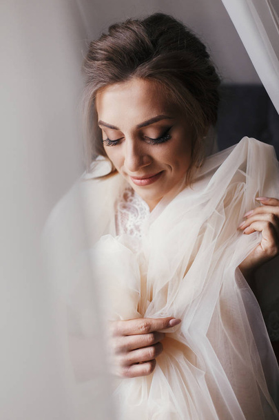 朝の柔らかな光で窓の近くポーズ シルクのローブで豪華な美しい花嫁。完璧なメイクのベールを押しながら笑顔で幸せなスタイリッシュな花嫁。テキストのためのスペース. - 写真・画像