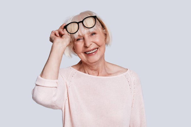wrinkled caucasian woman with short grey hair posing at grey background in studio and adjusting stylish glasses  - Foto, immagini