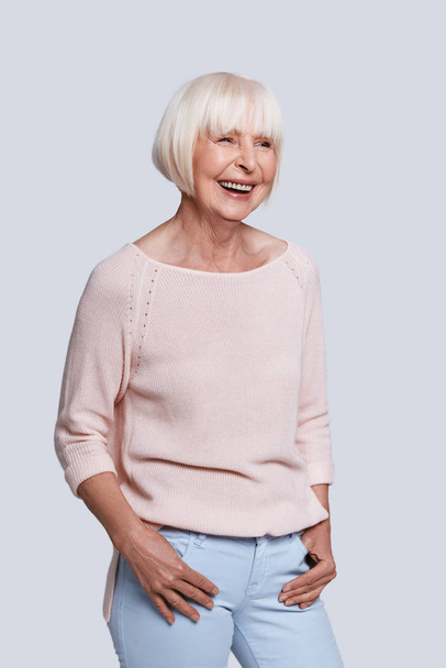 smiling caucasian woman in light sweater posing at grey background in studio with hands in pockets  - Photo, Image