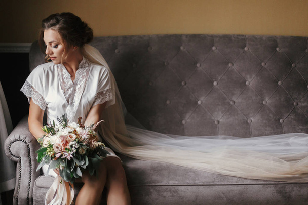 Gorgeous beautiful bride in silk robe and veil holding modern bouquet and sitting on stylish gray sofa. Happy stylish Bride with perfect makeup posing near window in soft light in the morning - Photo, Image