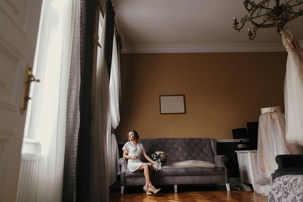 wunderschöne Braut in Seidengewand und Schleier mit modernem Strauß und sitzt auf einem stilvollen grauen Sofa. glückliche stilvolle Braut mit perfektem Make-up posiert am Fenster in sanftem Licht am Morgen - Foto, Bild