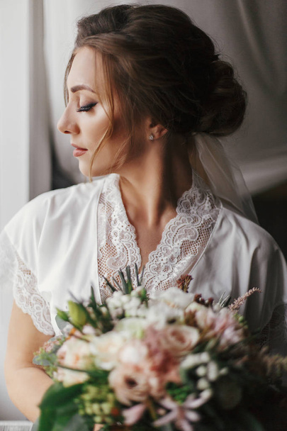 Gorgeous beautiful bride in silk robe and veil holding modern bouquet  posing in light. Happy stylish Bride with perfect hairstyle posing near window in the morning. Space for text. - 写真・画像