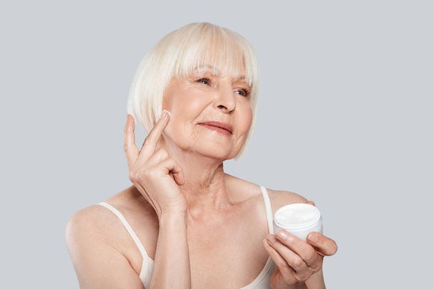 beautiful senior woman applying beauty cream while standing against grey background - Zdjęcie, obraz