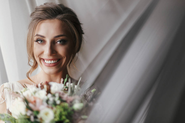 Gorgeous beautiful bride with bouquet posing at window in soft light. Happy Bride portrait in stylish gown, with perfect makeup, smiling and dreaming. Romantic moment. Space for text - Photo, Image