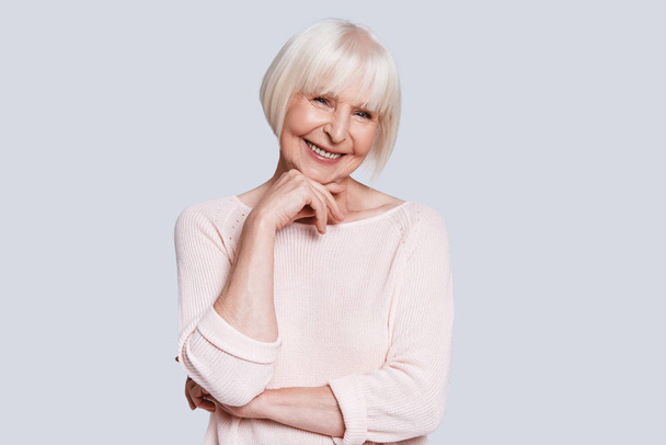 happy caucasian woman in light sweater and short grey hair posing at grey background in studio and holding hand at chin  - Photo, Image