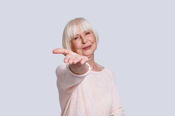 smiling caucasian woman in light sweater and short grey hair posing at grey background in studio, stretching arm to camera  - Fotó, kép