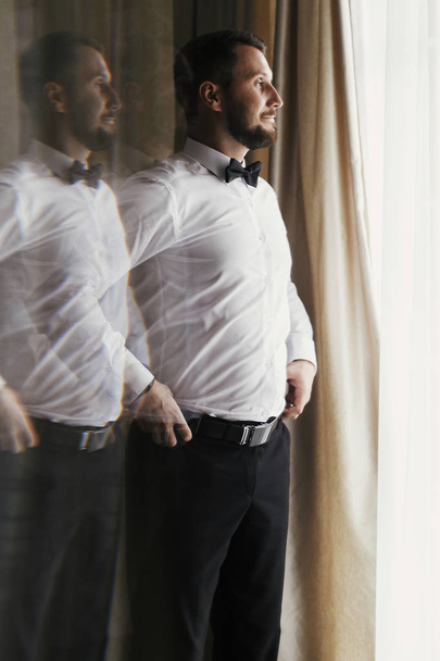 Stylish groom in white shirt and bow tie posing at window light. Confident and happy portrait of man. Groom getting ready in morning. Creative wedding photo with mirror reflection - Photo, Image