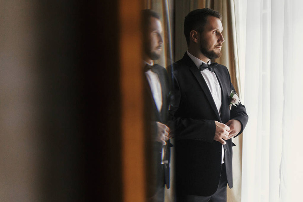 Stylish groom putting on suit and bow tie at window light. Confident and happy portrait of man. Groom getting ready in morning. Creative wedding photo with mirror reflection - Foto, Imagem