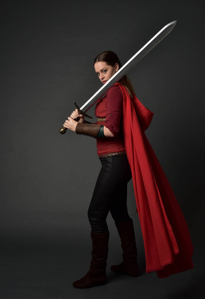 портрет дівчини брюнетки в червоному середньовічному костюмі і плащі. стояча поза, що тримає меч на сірому студійному фоні
. - Фото, зображення