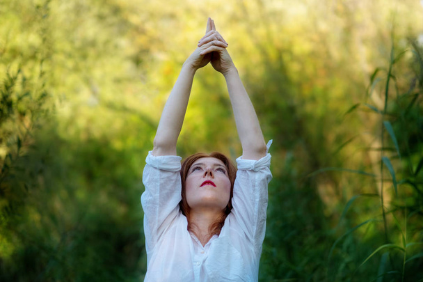 junge rothaarige Frau macht Yoga-Übungen mit erhobenen Händen in der Natur in Sportbekleidung in der Sonne in der Natur - Foto, Bild
