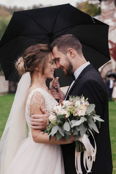 Gorgeous bride and stylish groom gently hugging under umbrella in rainy outdoors. Sensual wedding couple embracing. Romantic moments of newlyweds. Modern wedding photo - Photo, Image