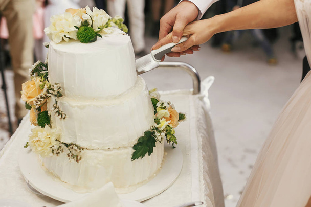 Gorgeous bride and stylish groom cutting together white wedding cake with roses at wedding reception. Happy wedding couple tasting cake. Romantic moments of newlyweds - Photo, Image