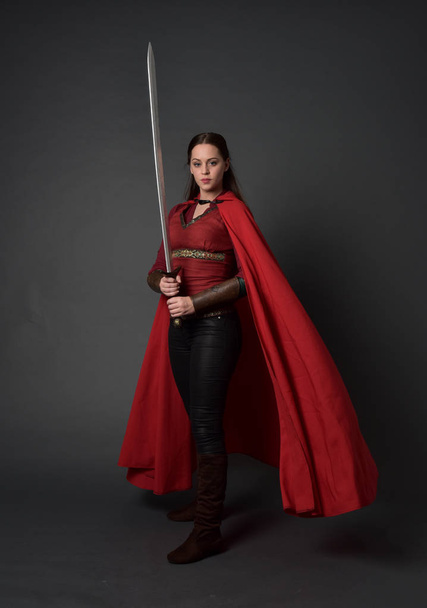 retrato de cuerpo entero de chica morena vestida con traje medieval rojo y capa. pose de pie sosteniendo una espada sobre fondo gris estudio
. - Foto, imagen