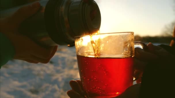 in winter forest man pours hot tea from thermos into glass goblet at sunset bright sun. girl holding glass of tea in hands of frosty evening in the park - Footage, Video
