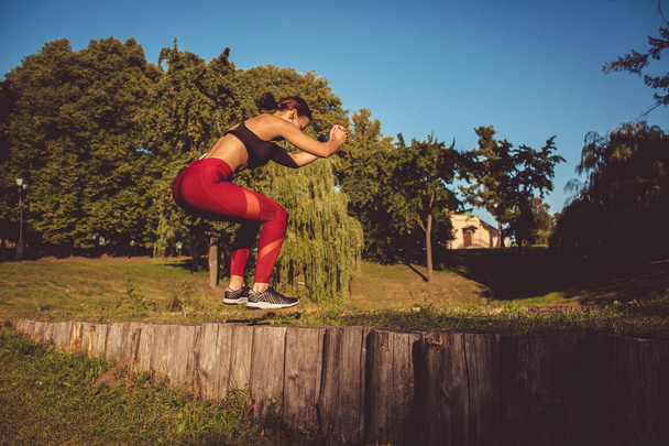 Mujer caucásica en leggins rojos y top deportivo calentándose en parque sobre postes de madera
 - Foto, Imagen