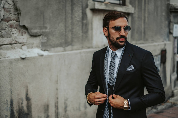 Bearded, elegant man buttoning suit while walking on the sidewalk in the city. - 写真・画像