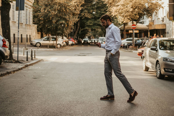 Homme beau négligent avec la main dans sa poche en regardant le téléphone portable tout en traversant la rue
 - Photo, image