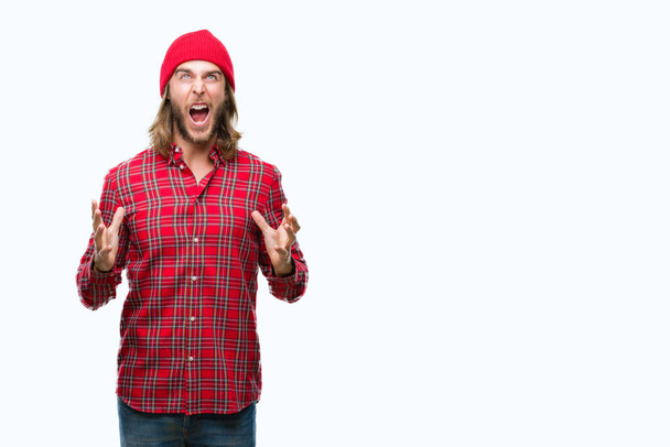 Young handsome man with long hair wearing red cap over isolated background crazy and mad shouting and yelling with aggressive expression and arms raised. Frustration concept. - Zdjęcie, obraz