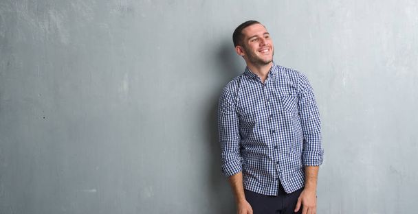 Молодий кавказький чоловік над сірою гранжевою стіною дивиться в сторону з посмішкою на обличчі, природним виразом. Сміється впевнено
. - Фото, зображення