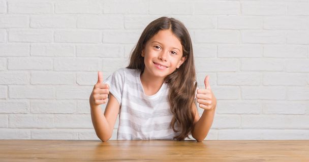 Young hispanic kid sitting on the table at home success sign doing positive gesture with hand, thumbs up smiling and happy. Looking at the camera with cheerful expression, winner gesture. - Photo, Image