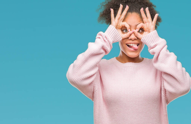 Mujer afroamericana joven que usa suéter de invierno sobre un fondo aislado haciendo un gesto aceptable como binoculares sacando la lengua, ojos mirando a través de los dedos. Expresión loca
. - Foto, imagen