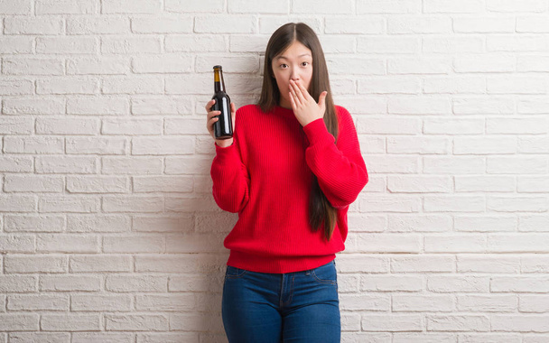 Jeune femme chinoise sur le mur de briques boire de la bière couvrir bouche avec la main choquée par la honte pour erreur, expression de la peur, peur dans le silence, concept secret
 - Photo, image
