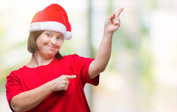 Mulher adulta jovem com síndrome de down usando chapéu de Natal sobre fundo isolado sorrindo e olhando para a câmera apontando com duas mãos e dedos para o lado
. - Foto, Imagem