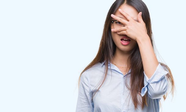 Jonge Aziatische zakenvrouw bril over geïsoleerde achtergrond gluren in schok voor gezicht en ogen met hand, op zoek door vingers met beschaamd expressie. - Foto, afbeelding