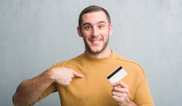 Молодий білий чоловік над сірою гранжевою стіною тримає кредитну картку з несподіваним обличчям, спрямованим пальцем до себе
 - Фото, зображення