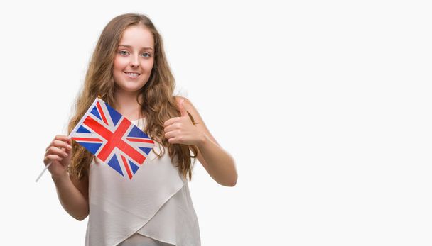 Молодая блондинка, держащая флаг Великобритании, счастлива с большой улыбкой, делая хороший знак, большой палец вверх пальцами, отличный знак
 - Фото, изображение