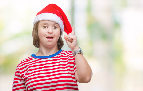 Mulher adulta jovem com síndrome de down usando chapéu de Natal sobre fundo isolado apontando dedo para cima com ideia bem sucedida. Saído e feliz. Número um.
. - Foto, Imagem