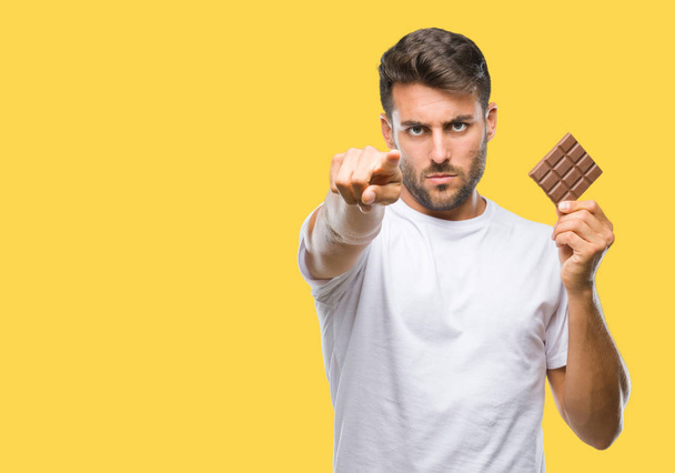 Młody przystojny mężczyzna jedzenie czekolady na białym tle wskazując palcem do aparatu i do Ciebie, ręcznie zarejestrować, gest pozytywny i pewnie z przodu - Zdjęcie, obraz