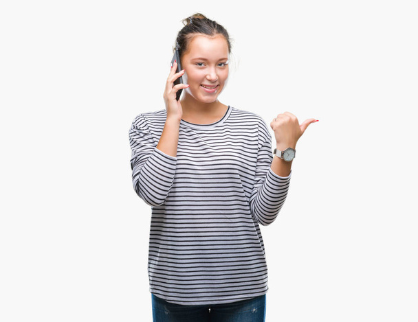 Jovem bela mulher caucasiana falando no smartphone sobre fundo isolado apontando e mostrando com o polegar até o lado com o rosto feliz sorrindo
 - Foto, Imagem