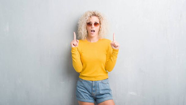 Young blonde woman with curly hair over grunge grey background amazed and surprised looking up and pointing with fingers and raised arms. - Foto, Bild